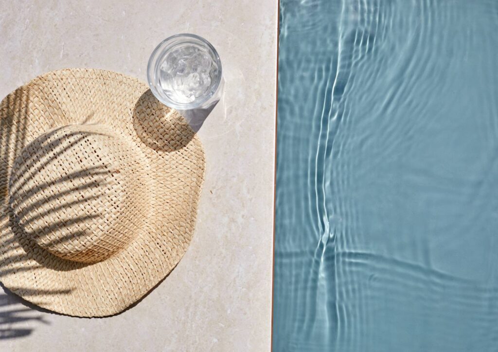 Sommerliche Stille – ein Strohhut und kühles Wasser auf warmem Stein neben einem türkisfarbenen Pool